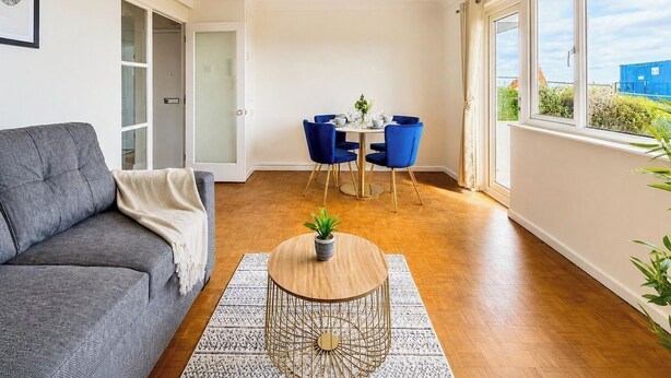 En moderne stue med en grå sofa, et rundt sofabord med en plante, et spiseområde med blå stole og et stort vindue med udsigt.