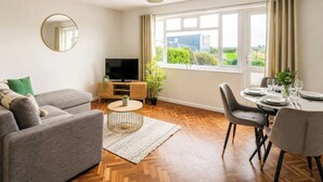 Sala de estar com sofá, televisão, mesa de jantar, cadeiras e uma janela grande com cortinas.