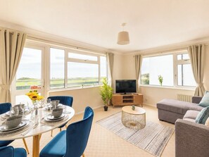 A modern living room with a dining table set, a sofa, a TV, and large windows with curtains.