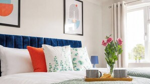 Una cama bien hecha con cabecera azul, almohadas blancas y verdes, un florero con flores y una bandeja con dos tazas y aperitivos.