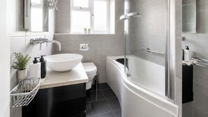 Un baño moderno con lavamanos, inodoro y bañera blancos, cabina de ducha con mampara de vidrio y una ventana que proporciona luz natural.