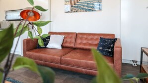 A living room with a brown leather sofa, a painting of a beach pier, and a plant with green leaves. - Cube Exclusive 6 (Nieuwvliet)