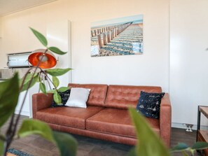 A living room with a brown leather sofa, a painting of a beach pier, and a plant with green leaves. - Cube Exclusive 6 (Nieuwvliet)