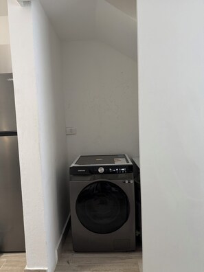 Ein kompakter Waschraum mit einer Samsung Waschmaschine und einem Trockner, einem Edelstahl-Kühlschrank und einem weiß gefliesten Boden.