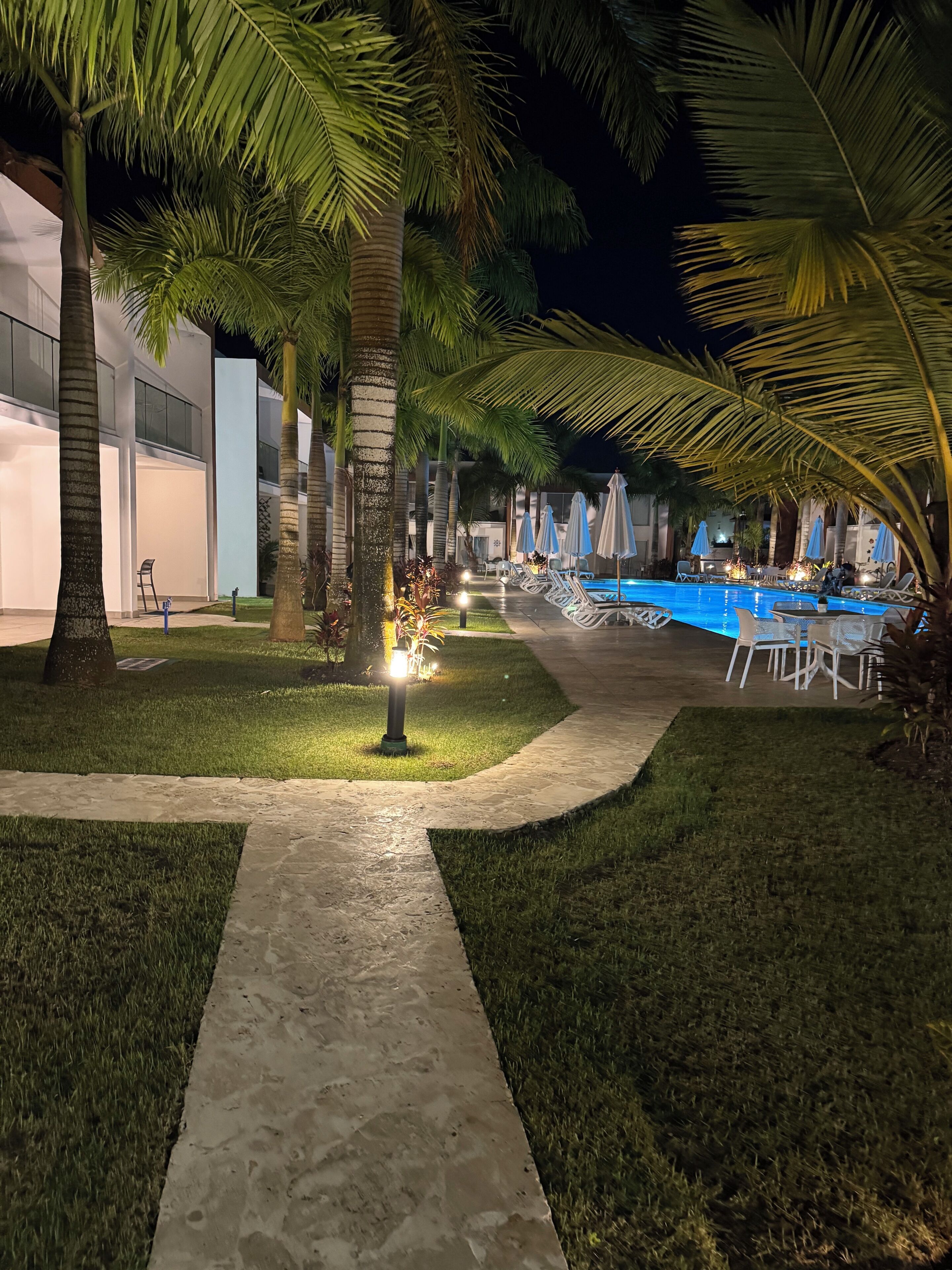 A nighttime outdoor pool area with lounge chairs and umbrellas, surrounded by palm trees and a pathway.