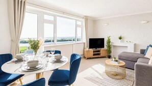 A modern living room with a dining area, large windows, a flat-screen TV, and a grey sofa.