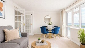 A modern living room with a sofa, a round coffee table, a dining table with chairs, a large window, and a view of the sea.