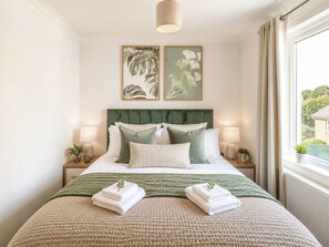 Een net opgemaakt bed met een groen hoofdeinde, twee nachtkastjes met lampen, en een raam met gordijnen.