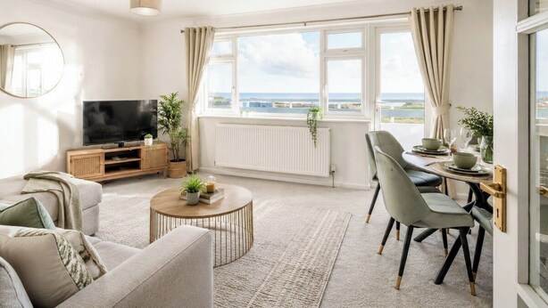 Una sala de estar moderna con televisor, sofá, mesa de comedor y sillas, ventanas amplias con vistas y una lámpara de techo.