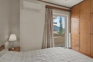 A bedroom with a bed, a nightstand with a lamp, a wardrobe, and a window with curtains. - Kons Beach Front Apartment with Sea View, Wi-Fi, and Air Conditioning (Kalamata)