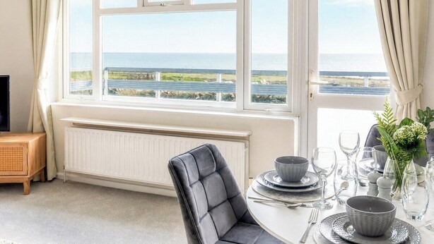 En matplats med ett bord dukat för fyra personer, en grå klädd stol och utsikt över havet genom stora fönster.