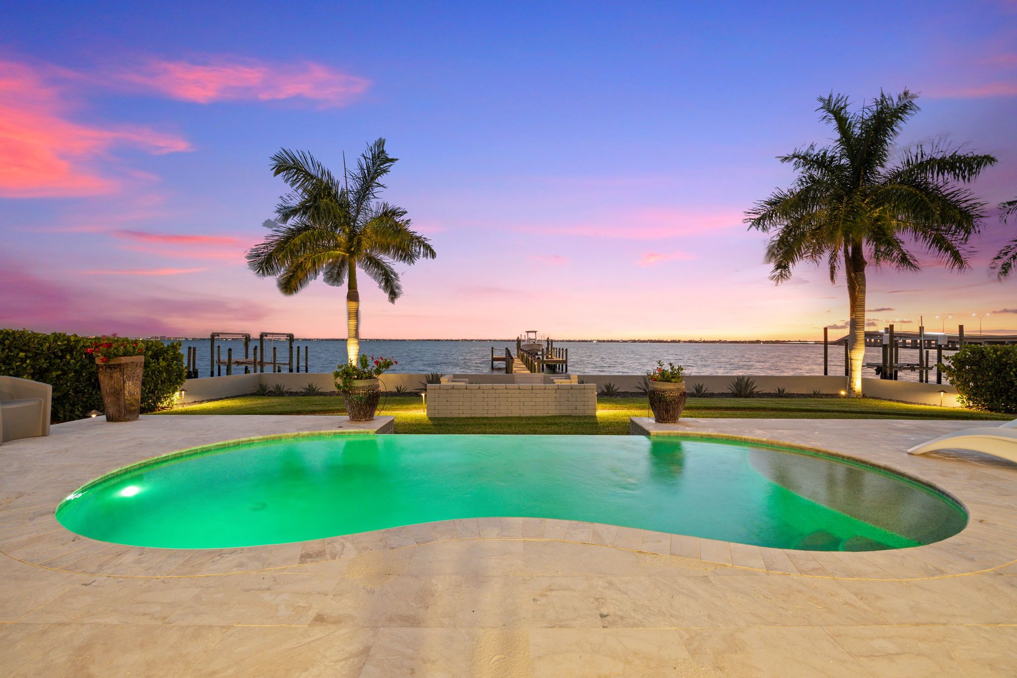 Piscina com borda curva, rodeada por um deck pavimentado, palmeiras e vista para o oceano ao pôr do sol.