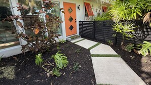Une maison moderne avec une porte orange distinctive, une clôture noire et un jardin où poussent différentes espèces de plantes.