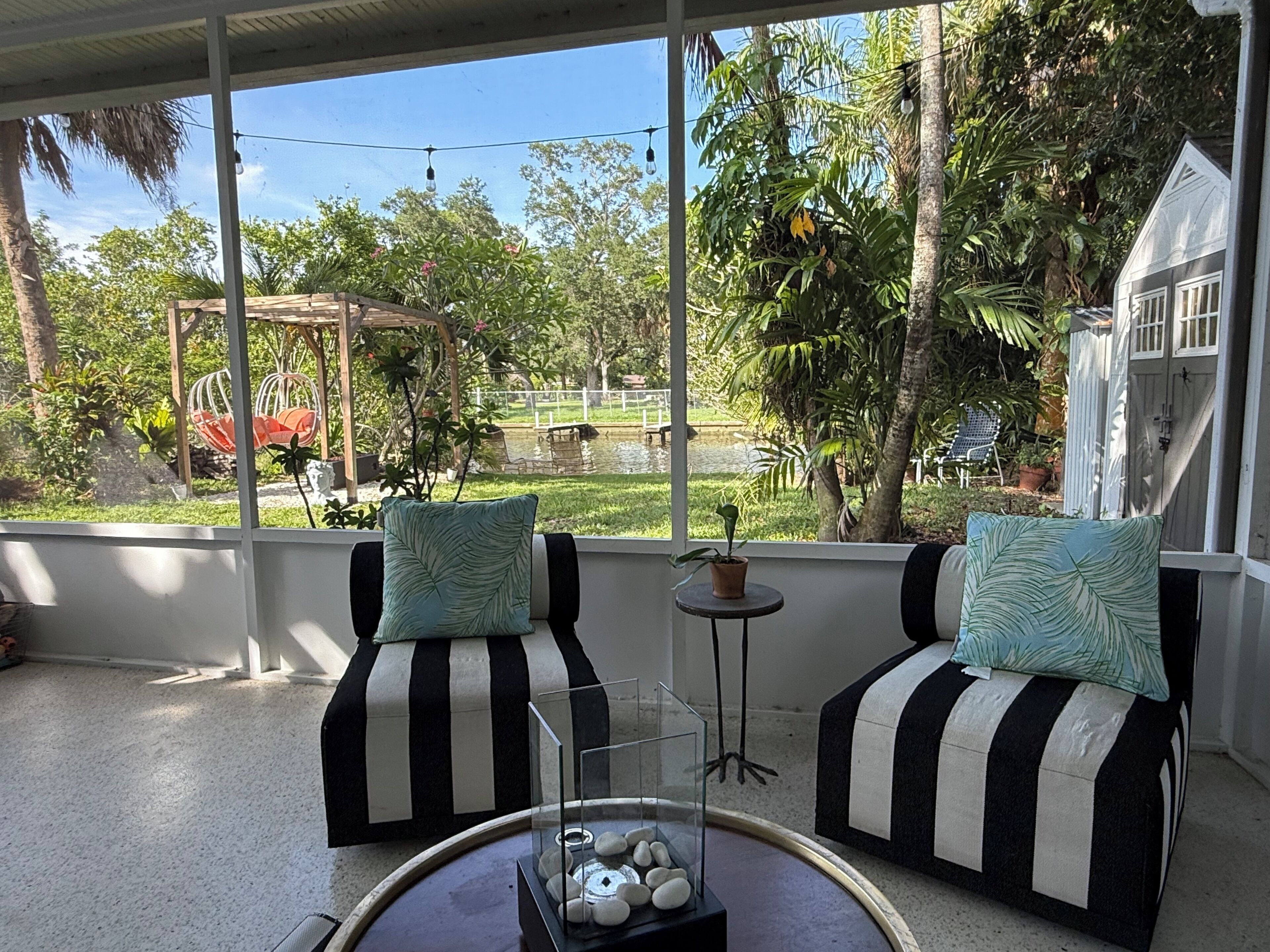 A room with two black and white striped sofas, a small table with a glass top, and a view of a garden with trees and a pond.
