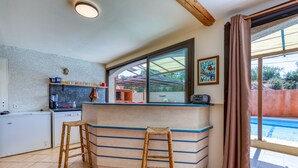 A kitchen with a bar area, two stools, a refrigerator, and a window overlooking a pool.