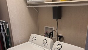 Un área de lavandería con dos lavadoras y secadoras de carga frontal, un estante con productos de limpieza y un enchufe eléctrico en la pared.