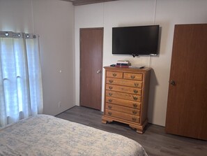 Une chambre à coucher avec un lit, une commode en bois, un téléviseur fixé au mur et une porte.