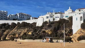 A coastal building with white walls and blue accents, situated on a sandy beach with a rocky hillside in the background. - Sunset View Albufeira 53 (Albufeira)