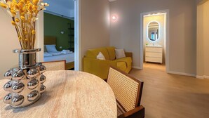 Quarto de hotel moderno com área de jantar, sofá amarelo, banheiro com espelho e cama com parede verde ao fundo.