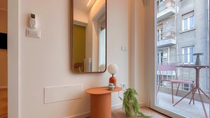 Um interior moderno com um espelho grande, uma mesa redonda pequena com uma planta e vista para uma varanda externa com cadeiras.