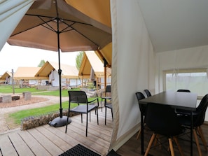 Ein zeltähnlicher Außenbereich mit Tisch, Stühlen, einem Grill und Blick auf andere Zelte.