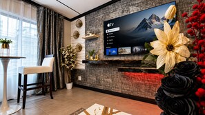 A modern living room with a flat-screen TV mounted on a textured wall, a dining area with a white table and chair, and decorative plants.
