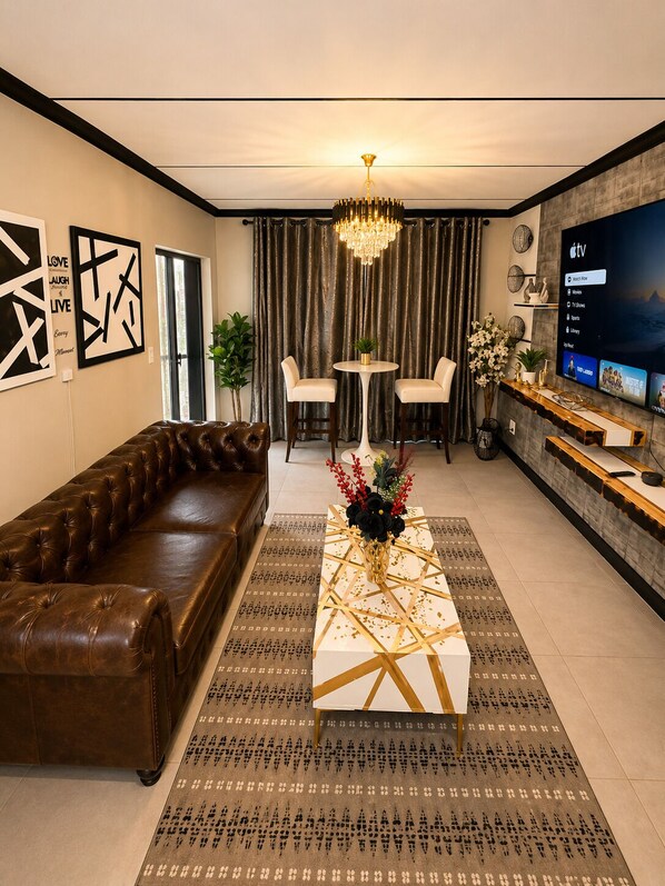 A modern living room with a leather sofa, a coffee table with a geometric design, a chandelier, and a flat-screen TV mounted on the wall.