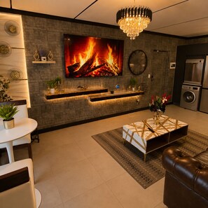 A modern living room with a fireplace, a television, a chandelier, and a kitchen area with a refrigerator.