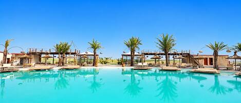 Una amplia piscina al aire libre con sillas de descanso y sombrillas, rodeada de palmeras y un cielo azul despejado.