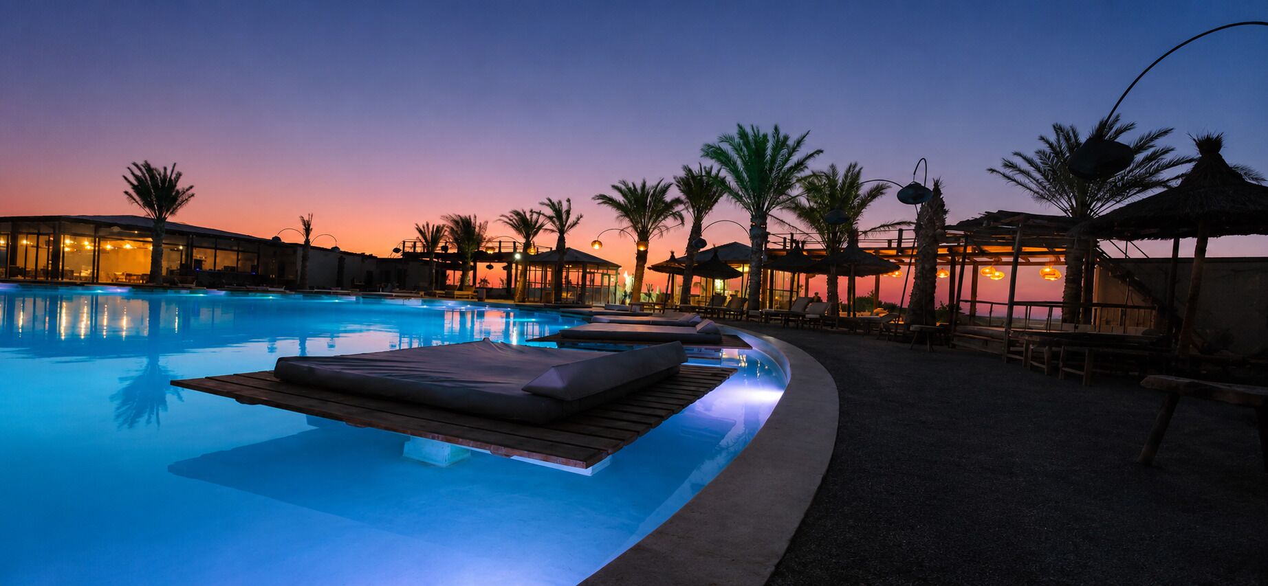 Piscina com espreguiçadeiras e guarda-sóis, rodeada por palmeiras e um pôr do sol.