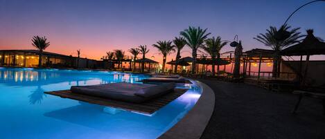 Piscina com espreguiçadeiras e guarda-sóis, rodeada por palmeiras e um pôr do sol.