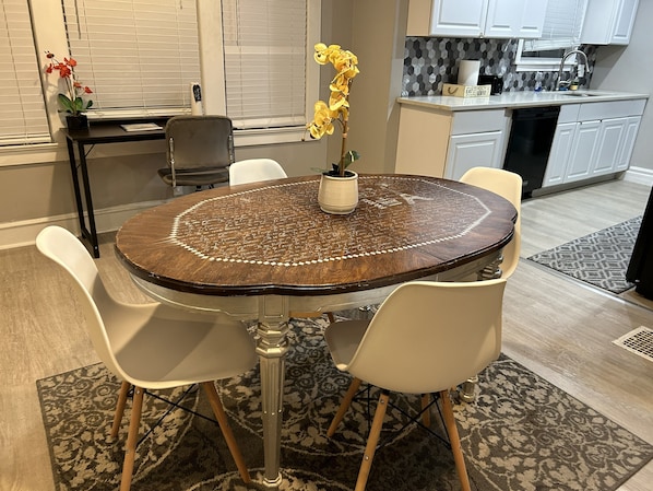 Un coin repas avec une table ronde, des chaises blanches, une plante en pot et une cuisine en arrière-plan.