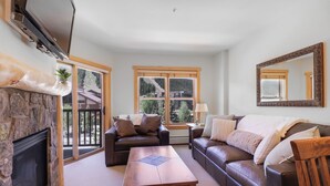 Ein Wohnzimmer mit Kamin aus Stein, zwei Sofas, einem Holztisch und einem großen Fenster mit Blick auf Bäume.