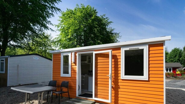 A small, single-story mobile home with an open door revealing a kitchen, a dining area with a table and chairs, and a small porch. - Holiday Home in Amstelveen With Terrace (Amstelveen)