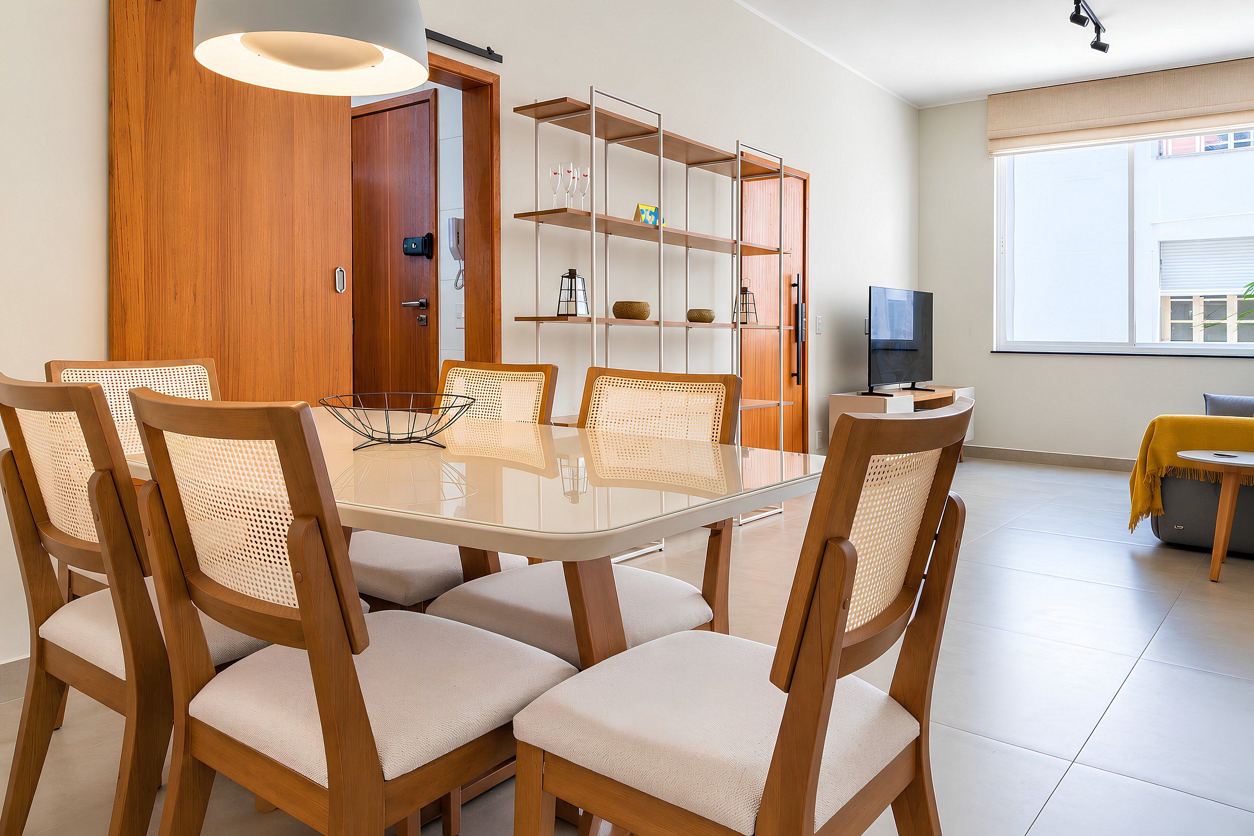 A dining area with a glass-top table, wooden chairs with woven seats, a bookshelf, and a television on a stand.