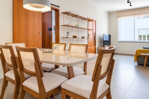 A dining area with a glass-top table, wooden chairs with woven seats, a bookshelf, and a television on a stand.