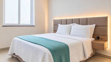 Una cama bien hecha con una colcha blanca y azul turquesa, una mesita de noche de madera con una lámpara, y una ventana con persianas.