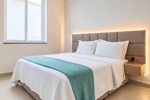 A neatly made bed with a white and teal blanket, a wooden nightstand with a lamp, and a window with blinds.