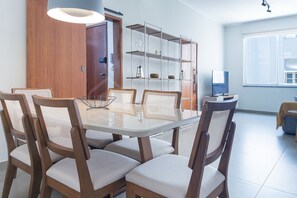 A dining area with a glass-top table, six wooden chairs with cushioned seats, a metal shelving unit, and a window with blinds.
