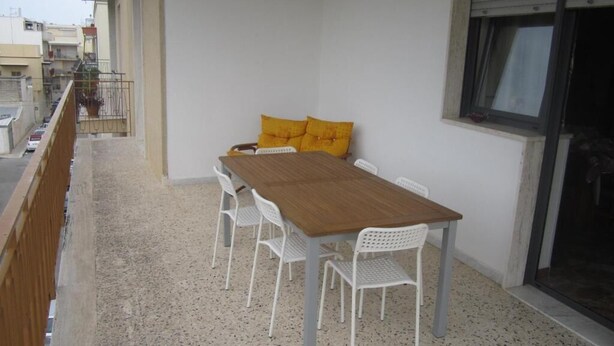 A balcony with a wooden table and white chairs, a window with blinds, and a view of the street. - Casa Raffaella (Polignano a Mare)