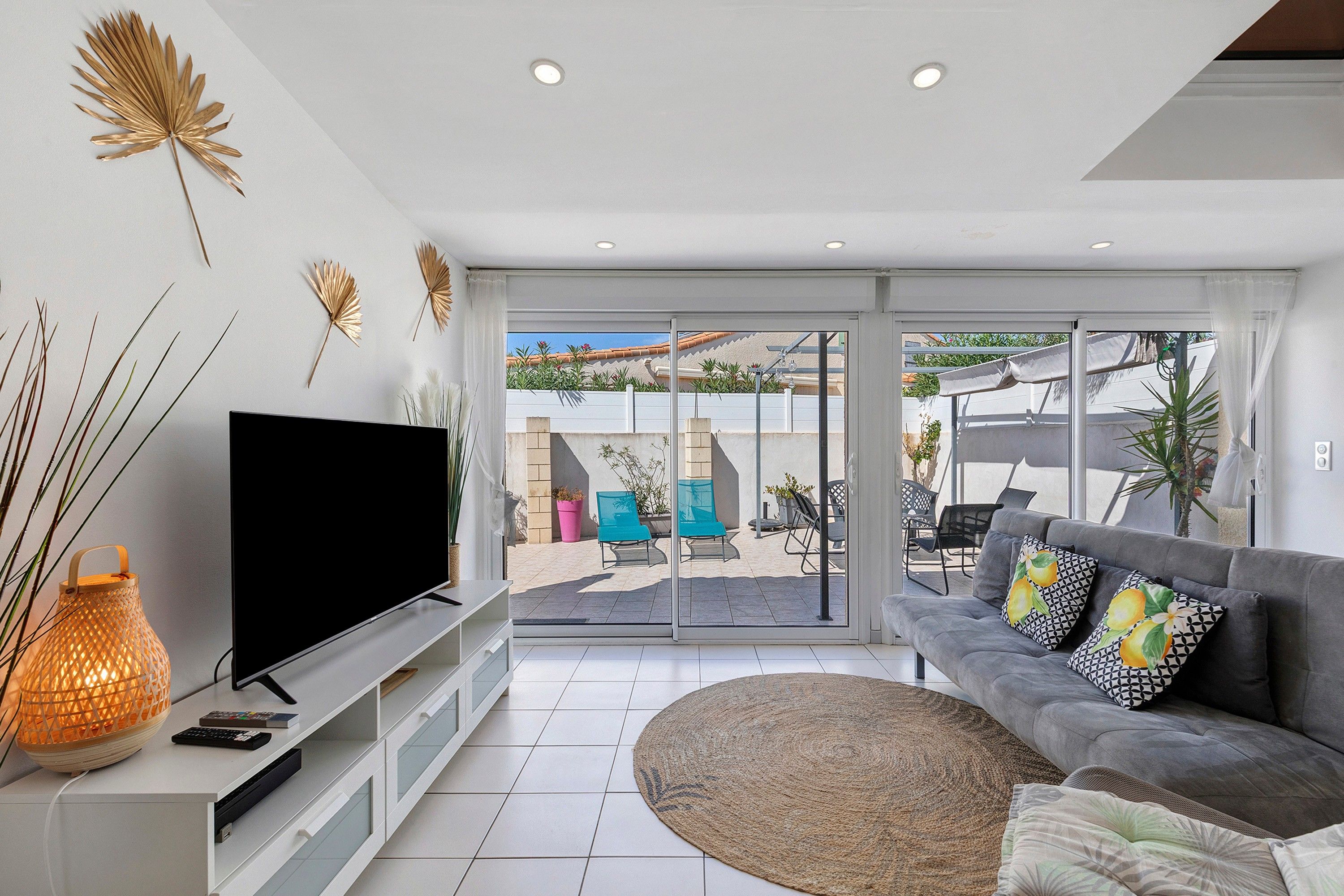 A modern living room with a flat-screen TV, a sofa with patterned cushions, and a large window overlooking a patio area.