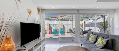 A modern living room with a flat-screen TV, a sofa with patterned cushions, and a large window overlooking a patio area.