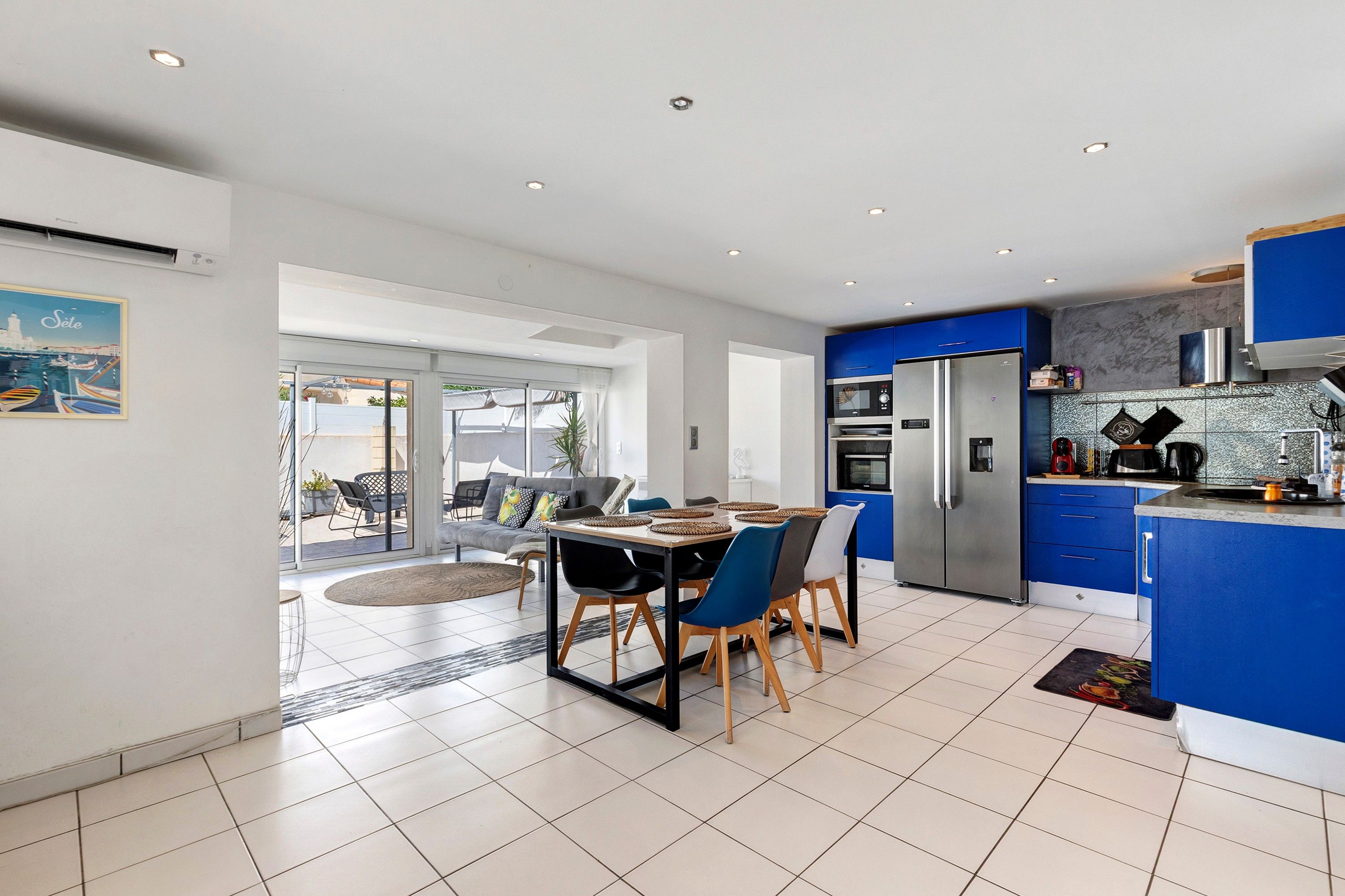 A modern kitchen with blue cabinets, a stainless steel refrigerator, and a dining area with a table and chairs.