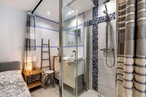 A modern bathroom with a glass shower enclosure, a small bedside table, and a bed with a patterned bedspread.