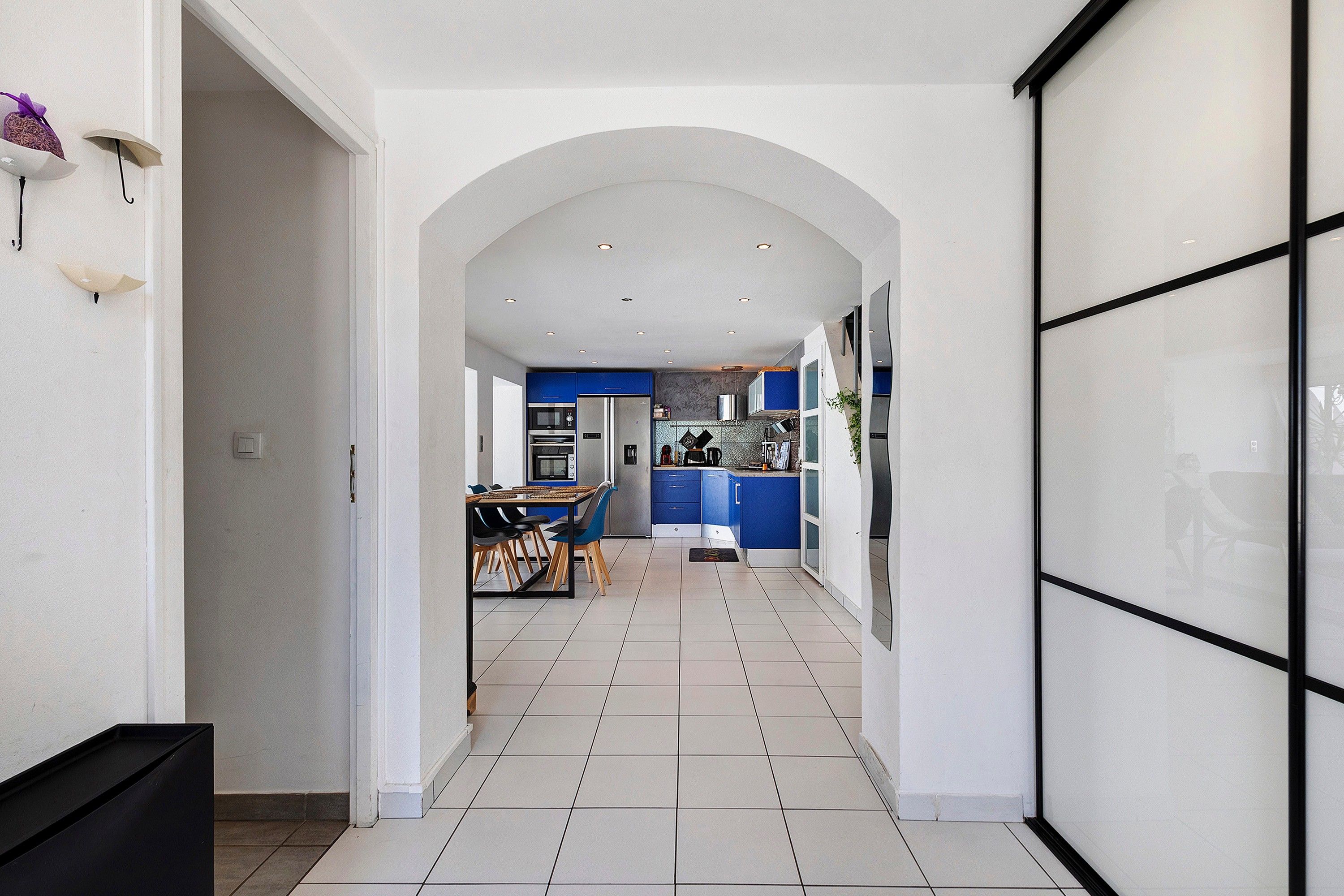 A modern kitchen with blue cabinets, a dining area with a table and chairs, and a hallway leading to a living area.