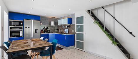 A modern kitchen with blue cabinets, a dining area with a wooden table and chairs, and a staircase with a plant on the side.