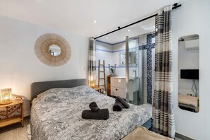 A modern bedroom with a large bed, a mirror, a bathroom with a glass door, and a TV.