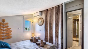 Une chambre à coucher avec un lit, une fenêtre, un miroir fixé au mur, et une salle de bain visible à travers une porte ouverte.