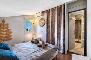 A bedroom with a bed, a window, a wall-mounted mirror, and a bathroom visible through an open door.