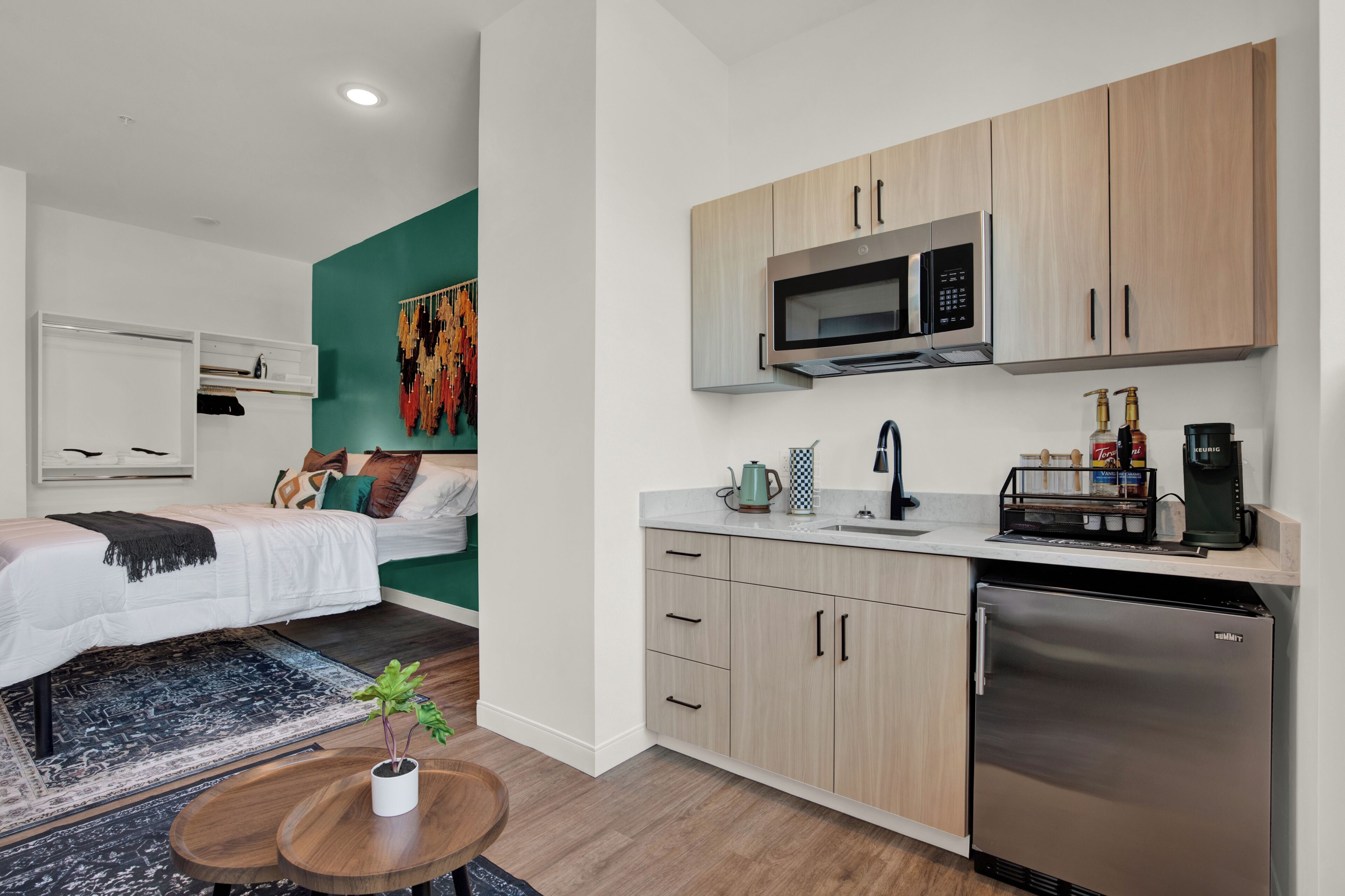 A modern room with a bed, a small table with a plant, a microwave, a coffee machine, and a refrigerator.